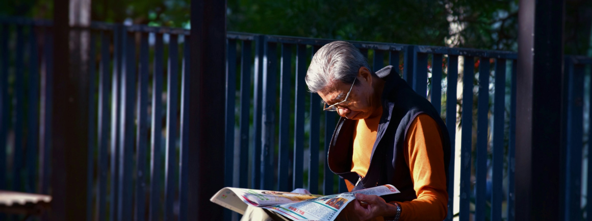 The Era of China's Senior-Citizen Economy Has Arrived, man reading newspaper while sitting on bench in front of wooden fence, photo by Mark Hang Fung So on Unsplash